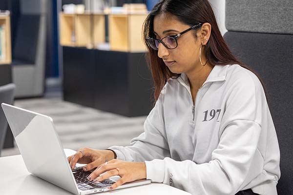 Uxbridge student sitting down at a desk and working on a laptop. She is wearing black glasses and a light coloured jumper.